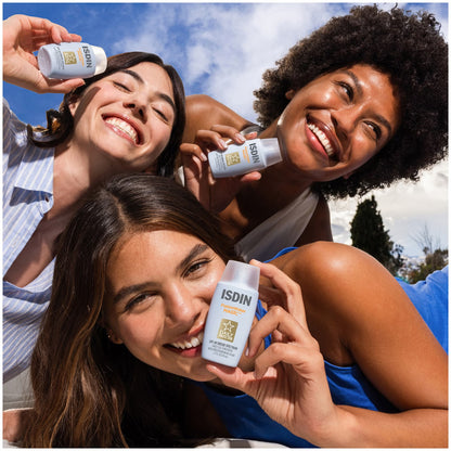 Three women holding ISDIN sunscreen bottles outdoors with a clear blue sky.