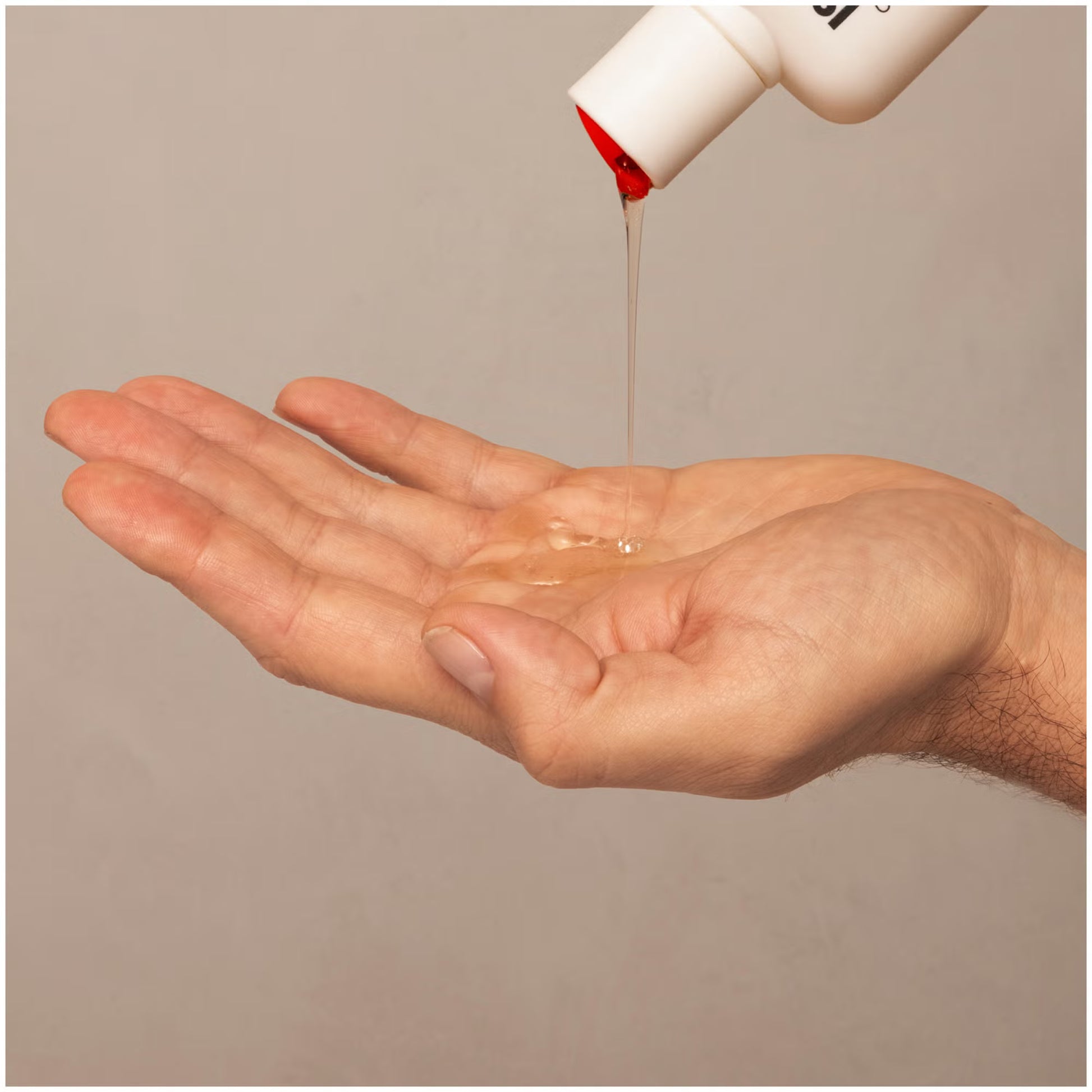 Hand pouring a clear liquid from a white bottle with red cap onto a beige background