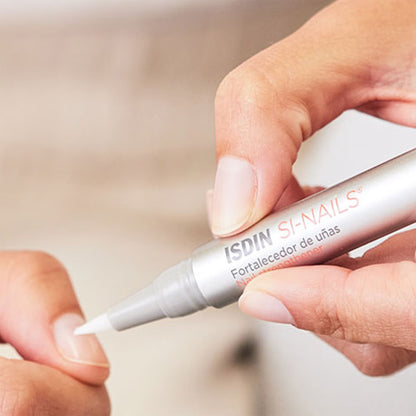 Hand applying a nail strengthener pen labeled 'ISDIN SI-NAILES' to a fingernail.