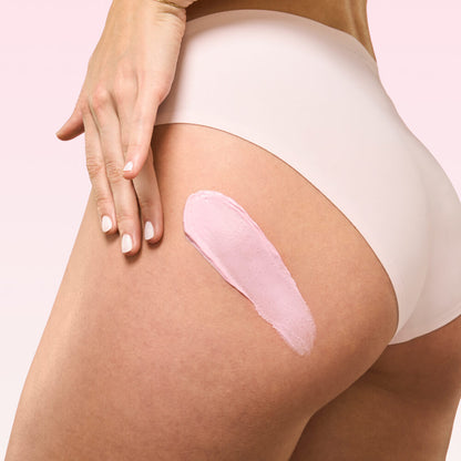 Person applying a pink cream or lotion to their leg on a light pink background