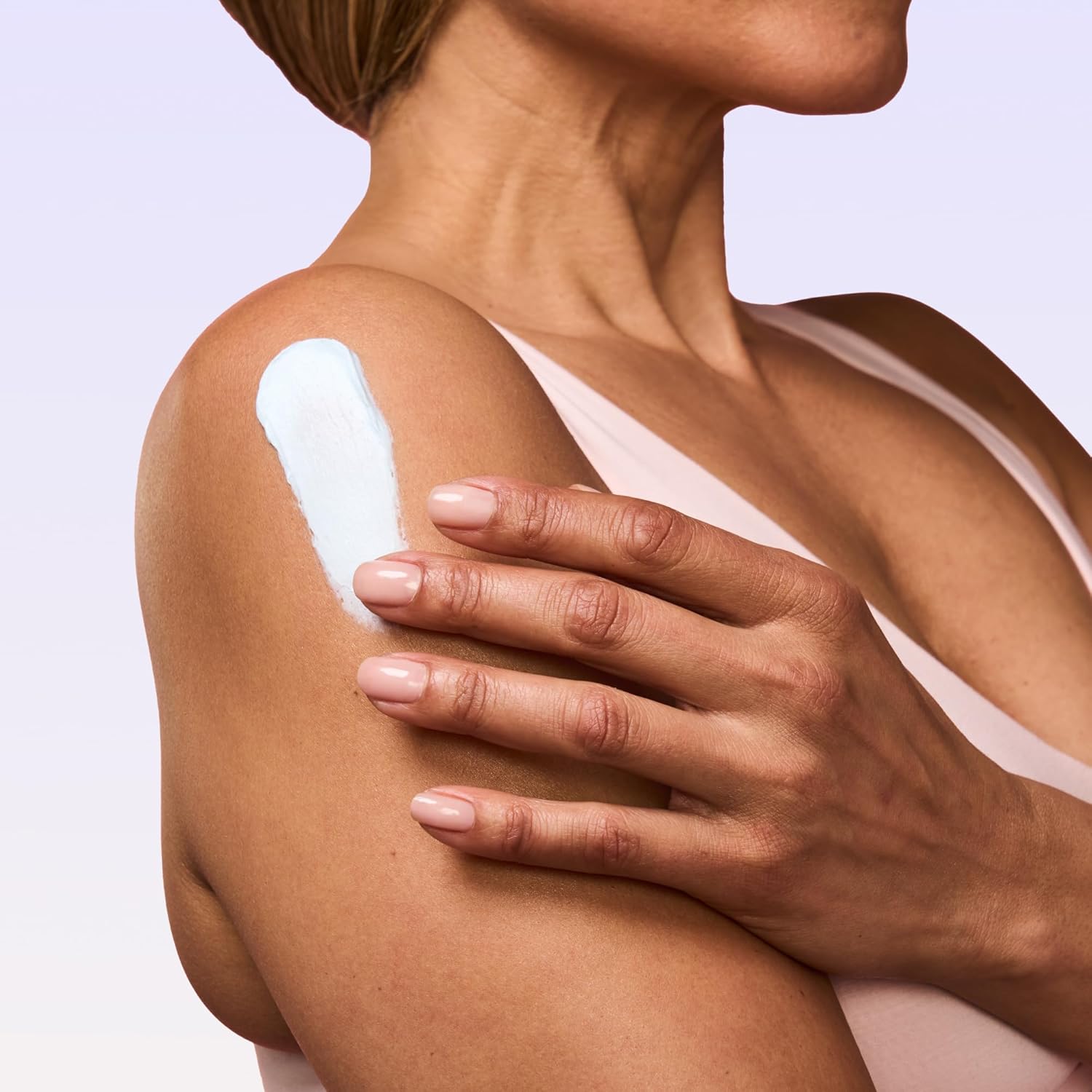 Person applying a white cream or lotion to their shoulder against a light purple background